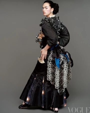 Fashion editorial shot: a woman in an elaborate black dress poses in a studio with a grey background, holding a gold fan and facing the camera.