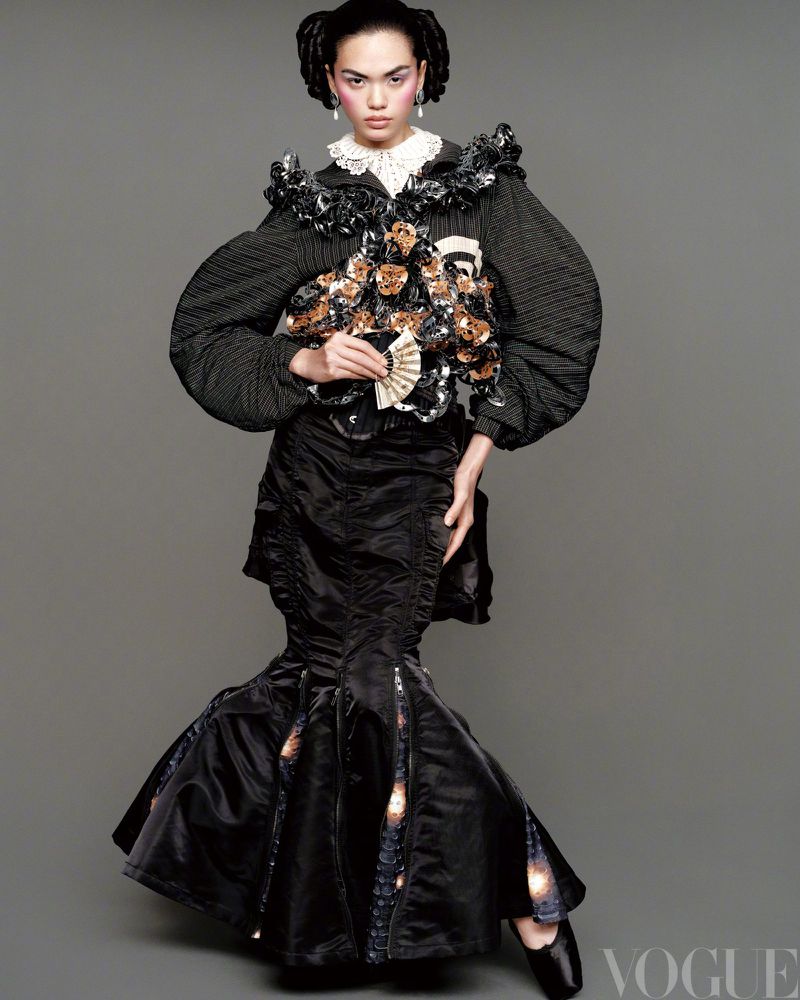 A person poses in an elaborate black gown with floral embellishments and a white lace collar against a neutral backdrop in a studio shot.