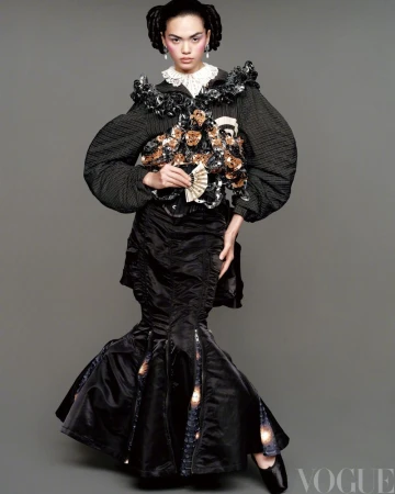 A person poses in an elaborate black gown with floral embellishments and a white lace collar against a neutral backdrop in a studio shot.