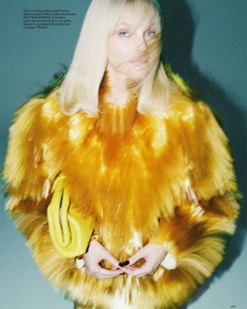 Model Jessica Stam wearing a voluminous textured golden-yellow fringe top and holding a yellow clutch against a teal studio backdrop.