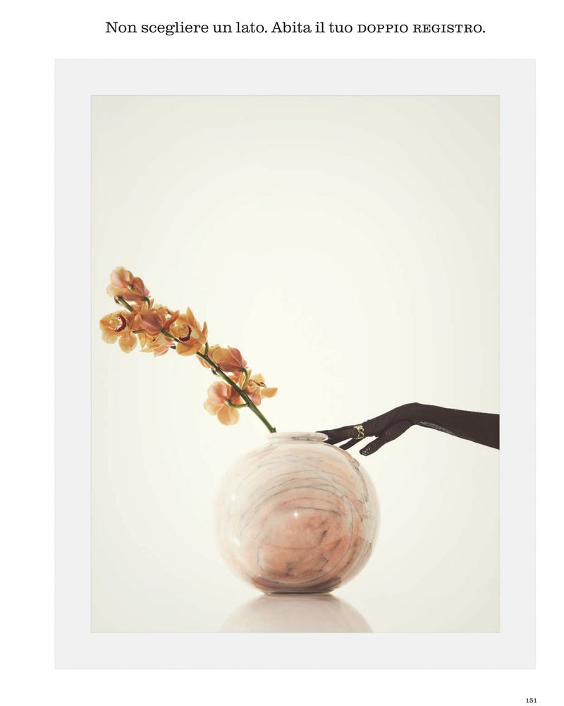 Round glass vase with an orange orchid branch beside a black gloved hand wearing a gold ring on a white background.