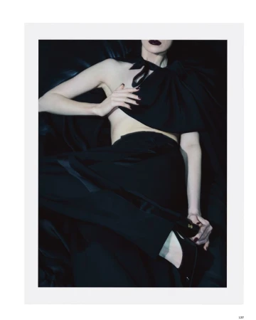 Jessica Stam posing in a black asymmetrical draped top, black trousers, and pointed high heels against a dark backdrop.