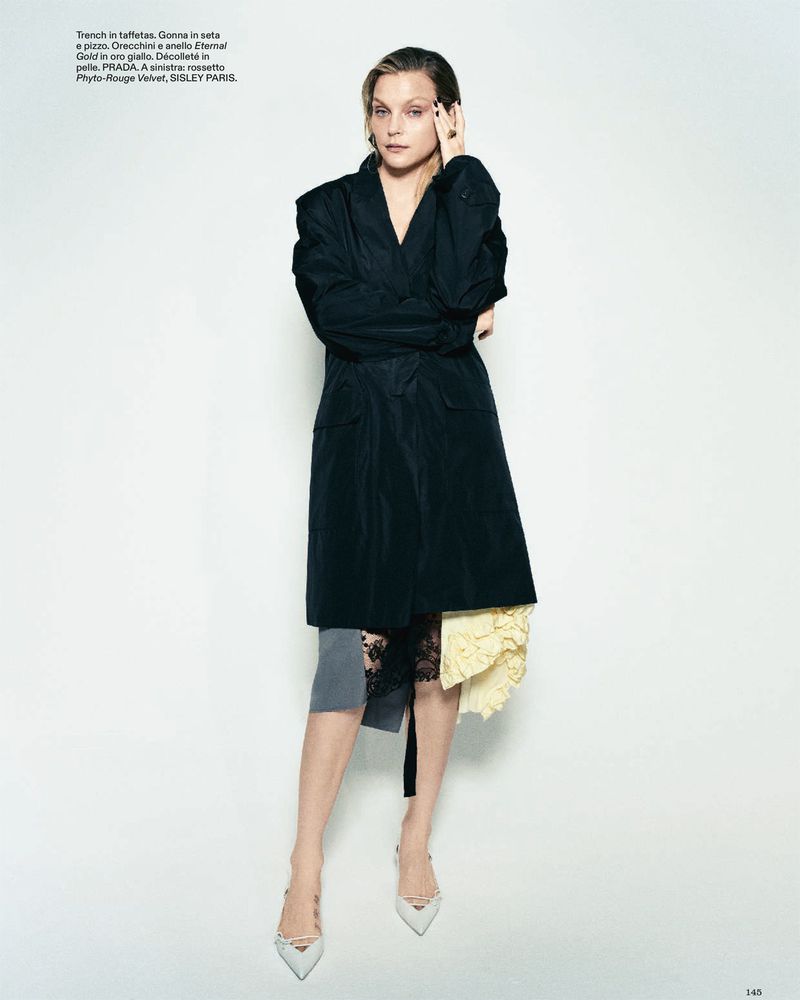 Model Jessica Stam poses in a black trench coat over a lace skirt and pointed heels against a white studio background.