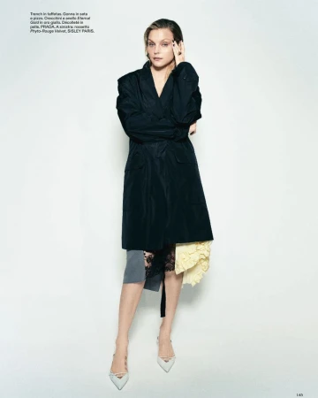 Model Jessica Stam poses in a black trench coat over a lace skirt and pointed heels against a white studio background.