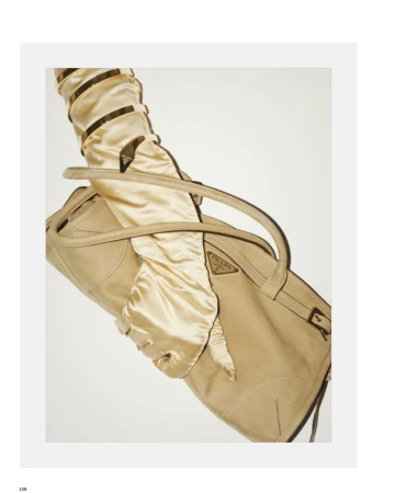 Arm in a champagne satin glove and gold cuff bracelet holding a beige designer handbag against a white background.
