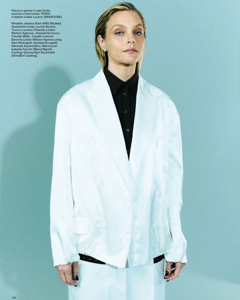 Model Jessica Stam wearing an oversized white blazer over a black button-down shirt against a pale teal studio backdrop.