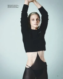 Model Jessica Stam wearing a black cropped long-sleeve sweater and high-cut sheer tights with arms raised against a grey backdrop.