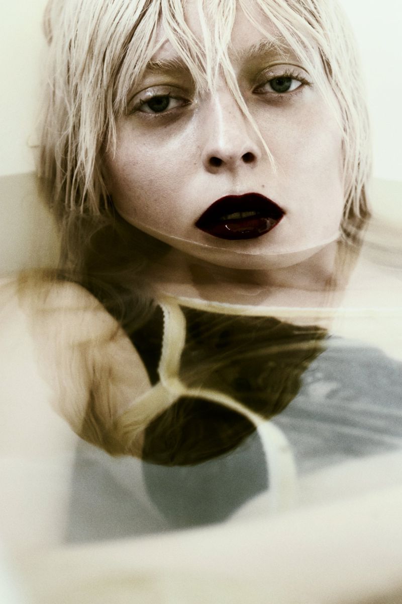 Close-up portrait of a woman with wet blonde hair and dark plum lipstick submerged in water inside a bathtub.