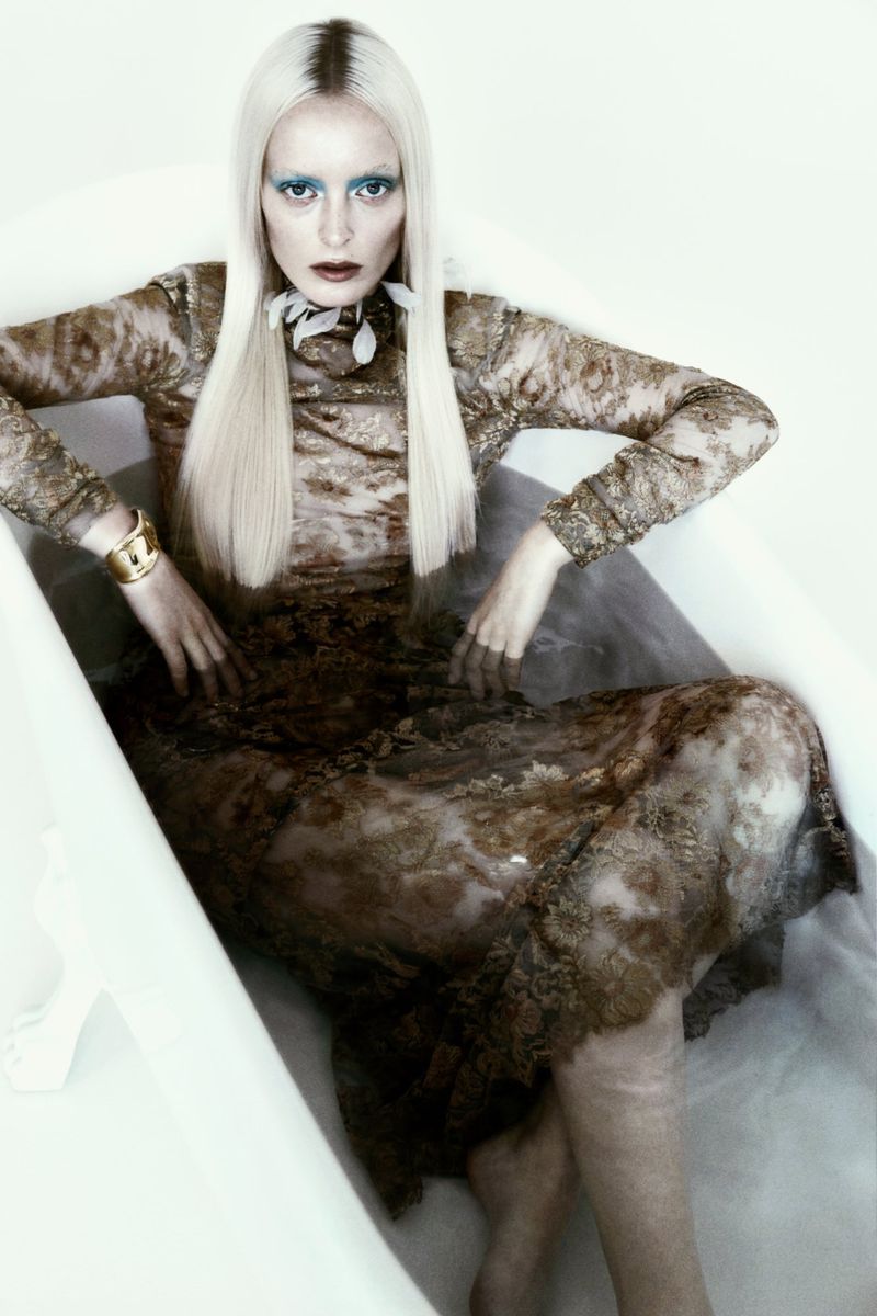 Blonde woman wearing a brown sheer lace dress lying submerged in a white bathtub for Numero Netherlands.