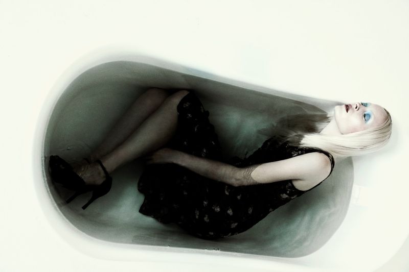 Blonde woman in a black dress and high heels lying curled inside an empty white bathtub from an overhead perspective.