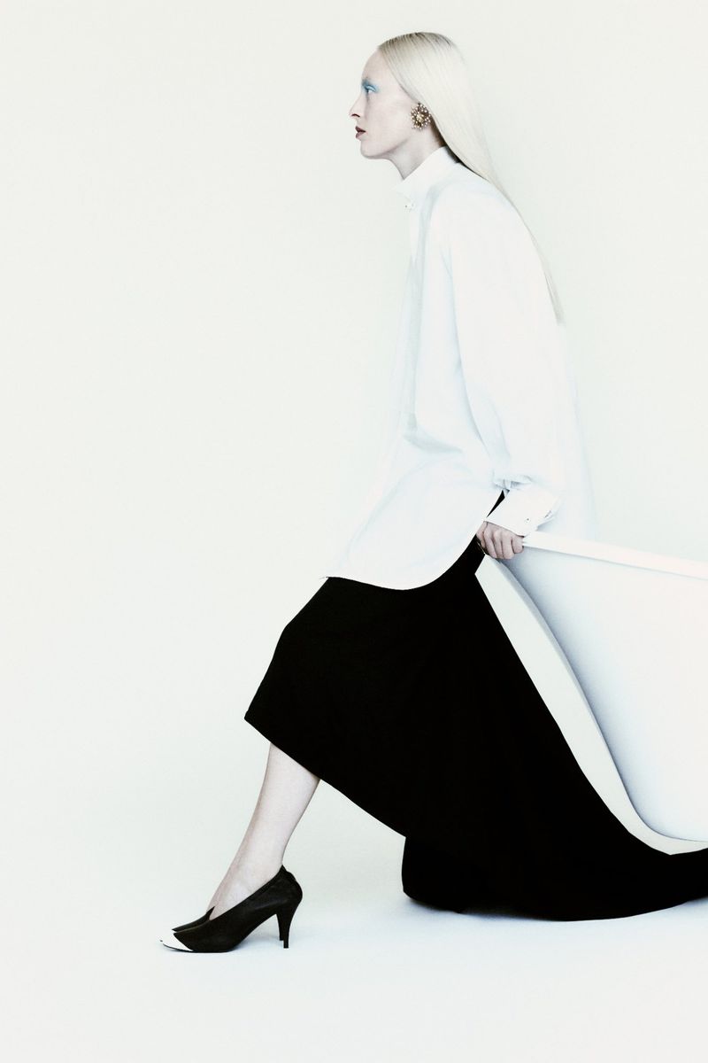 Fashion model in profile wearing an oversized white button-down shirt and long black skirt against a white studio background.