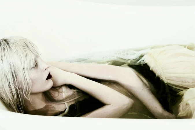 Model with platinum blonde wet hair submerged in a white bathtub wearing a pale ruffled tulle dress.