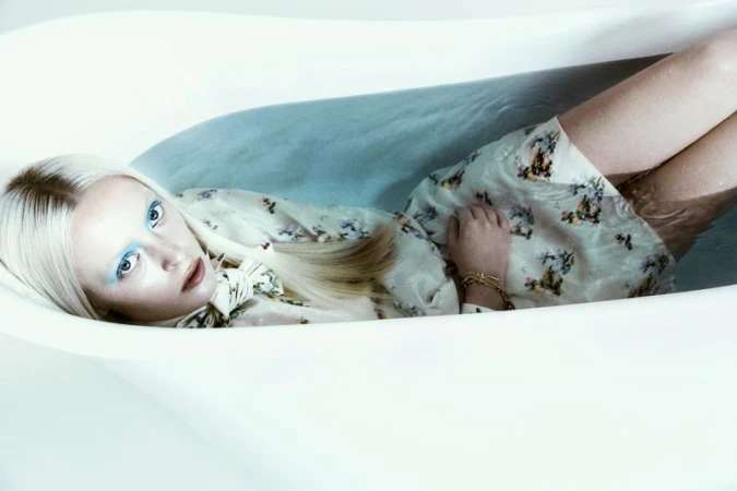 Blonde woman with blue eyeshadow lying in a floral dress inside a white bathtub filled with water.