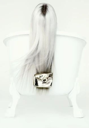 Long platinum blonde hair cascading over the back of a white clawfoot bathtub with a small metallic quilted purse attached.