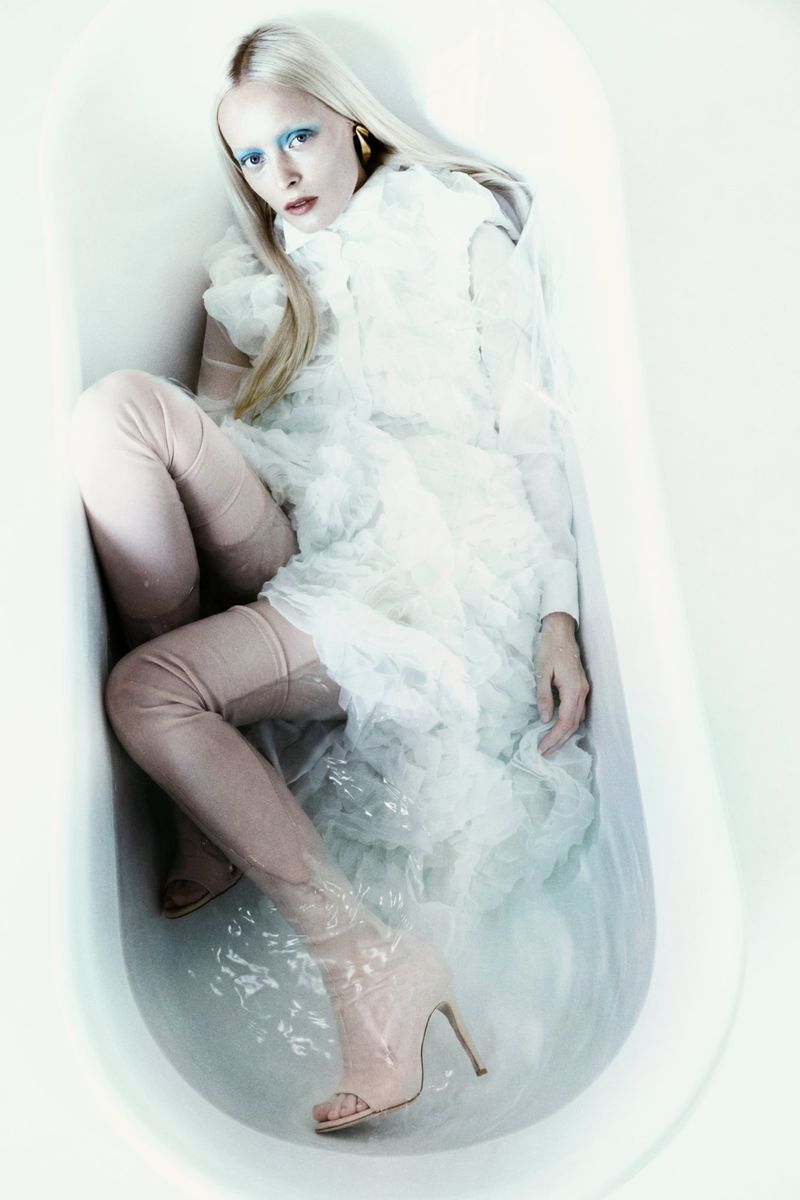 Blonde woman in a white ruffled dress and translucent thigh-high heeled boots lying in a water-filled bathtub with blue eye makeup.