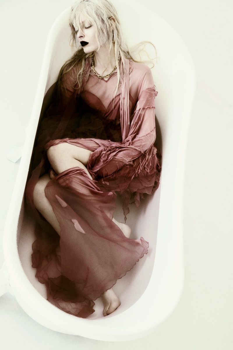 Model with long blonde hair and dark lipstick reclining inside a white bathtub wearing a sheer mauve dress