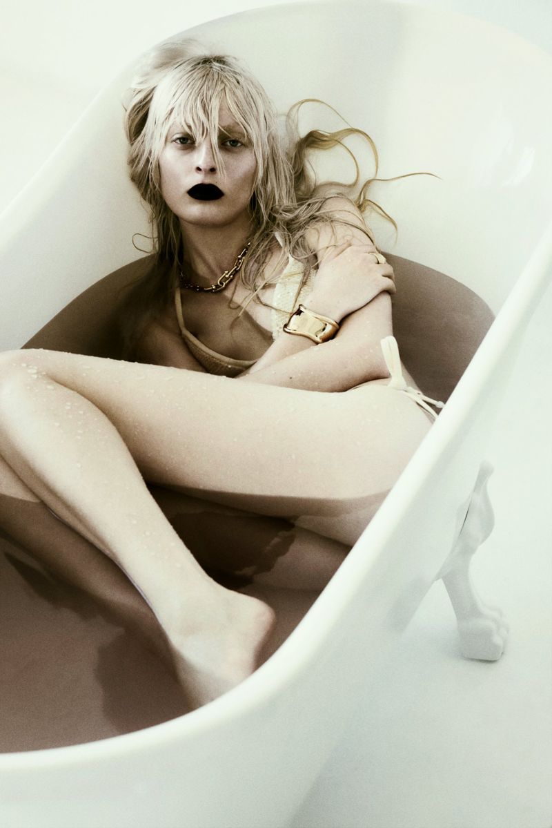 Blonde woman wearing dark lipstick and gold jewelry reclining inside a white clawfoot bathtub in a studio setting.