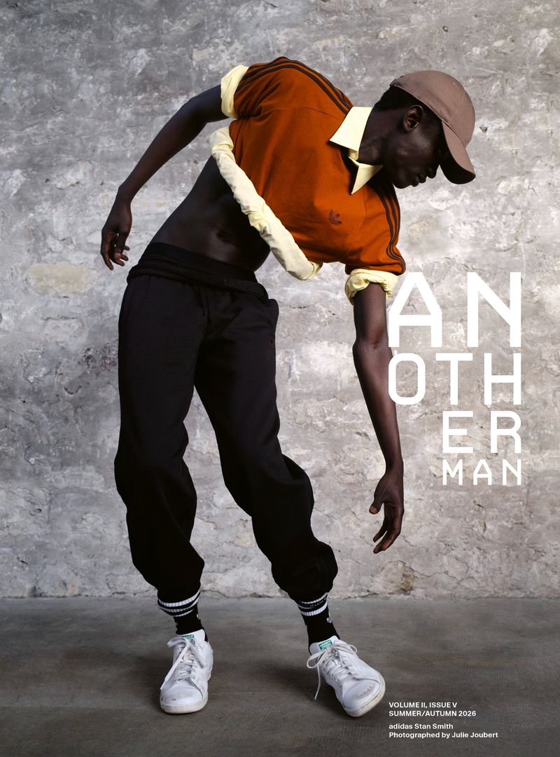 Black man wearing a rust brown cropped polo shirt and black trousers leaning forward against a rough stone wall background