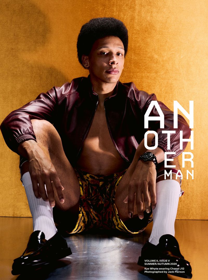 Kye Whyte seated on a reflective floor in an unzipped maroon leather jacket, patterned shorts, white socks, and black loafers.