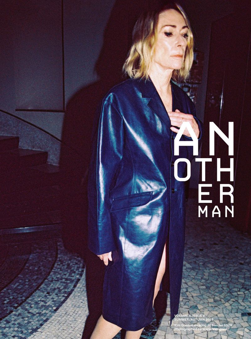 Kim Gordon wearing a dark leather coat standing in an interior space with a tiled floor and curved staircase.