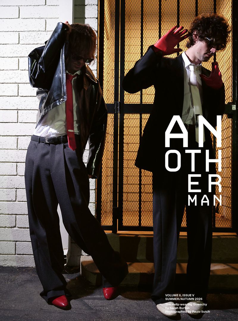 Two men in dark suits and red gloves posing outside against a brick wall and a metal security gate for AnOther Man Magazine.