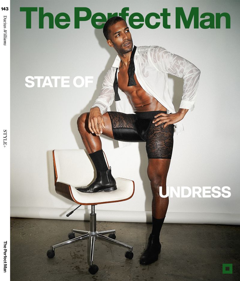 A male fashion model poses indoors in unbuttoned shirt, lace shorts, and boots next to a white chair for a magazine cover shoot.