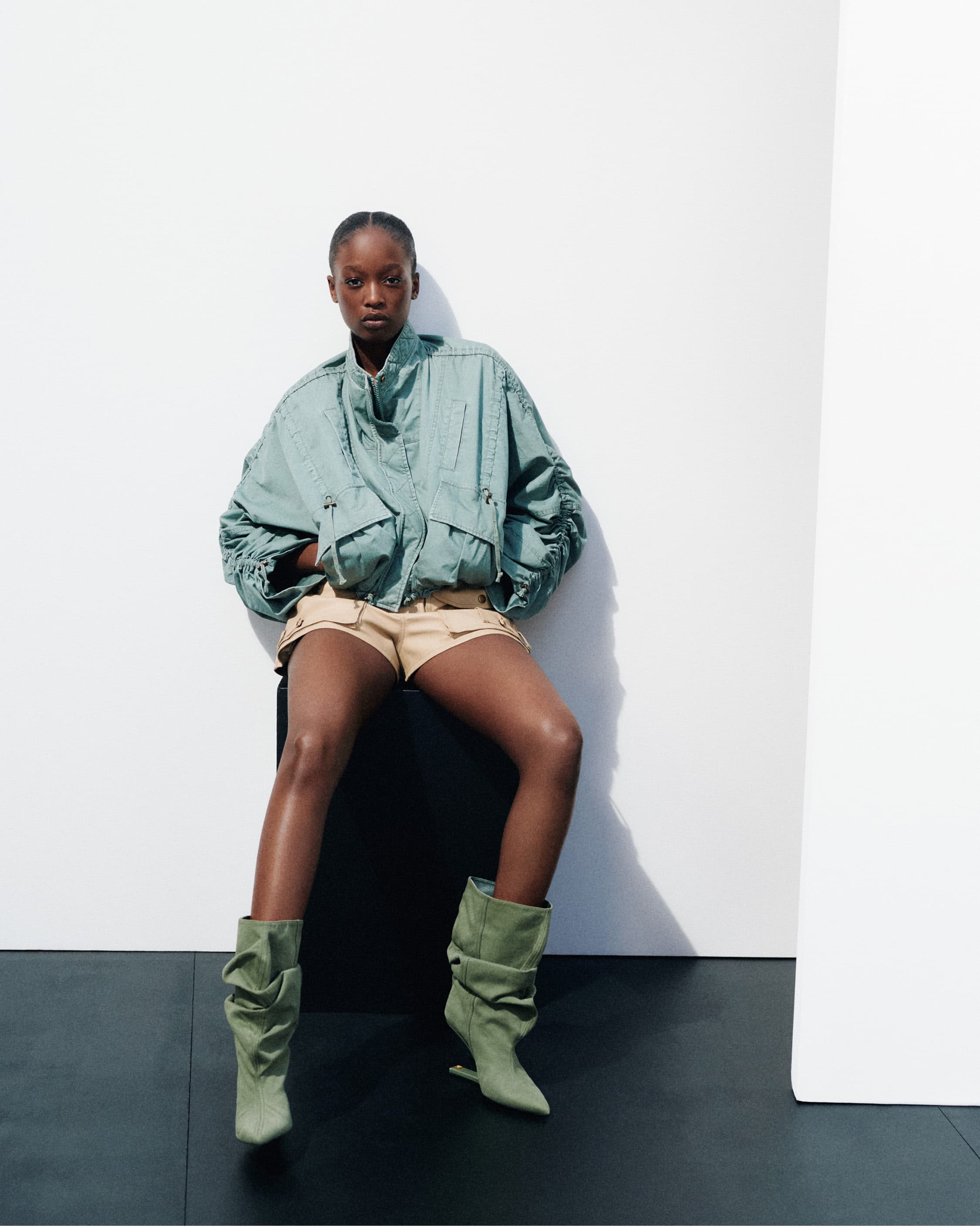 Model wearing a sage green jacket, beige shorts, and matching slouchy boots seated against a plain white wall.