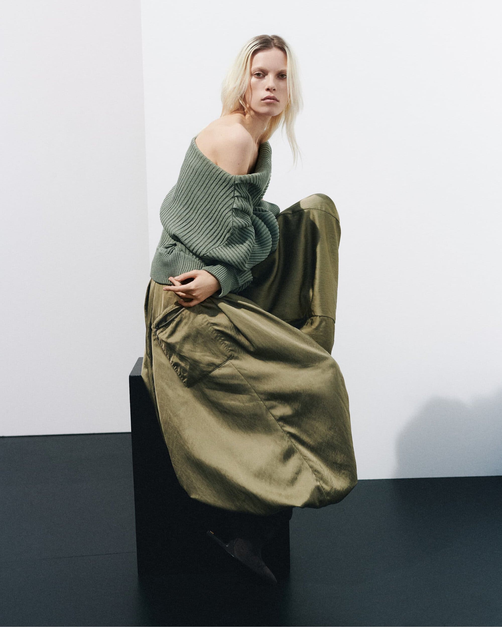 Model wearing an olive green off-the-shoulder ribbed sweater and wide trousers seated on a dark block in a white studio.