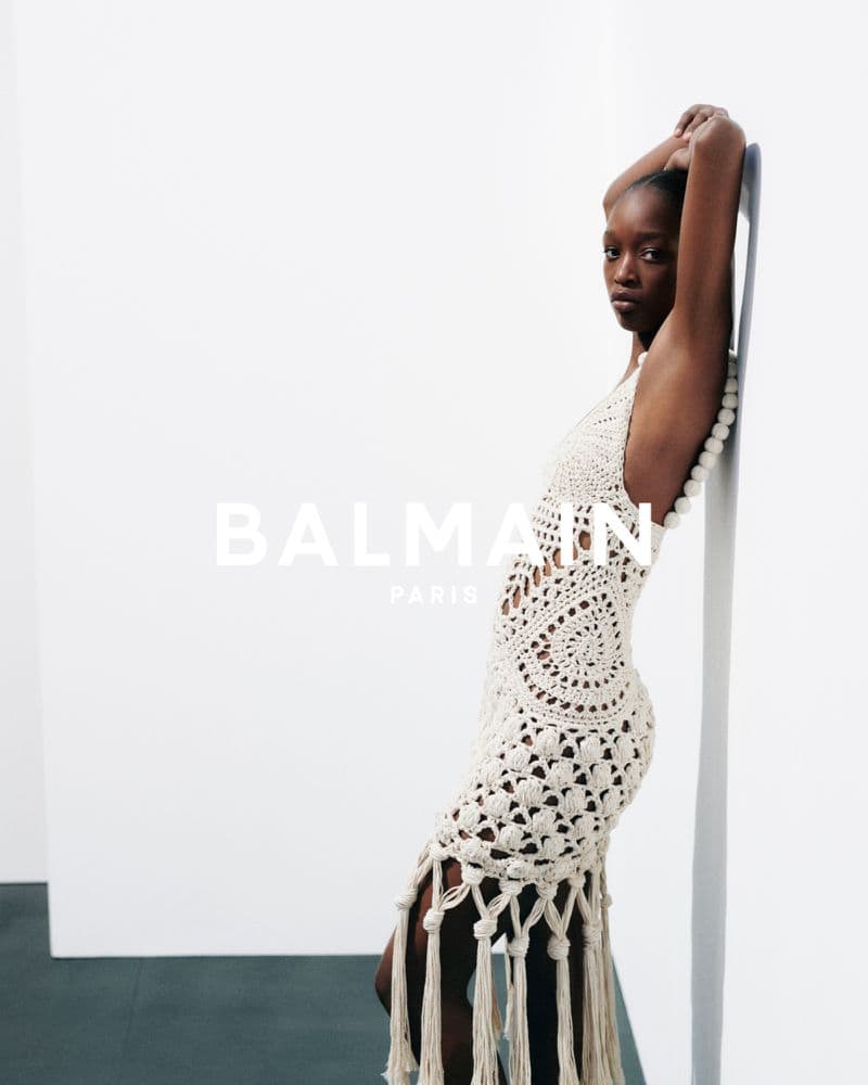 Black model leaning against white wall wearing a cream crocheted fringed dress for Balmain Spring 2026 campaign.