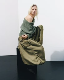 Model wearing an olive green off-the-shoulder ribbed sweater and wide trousers seated on a dark block in a white studio.
