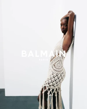 Black model leaning against white wall wearing a cream crocheted fringed dress for Balmain Spring 2026 campaign.