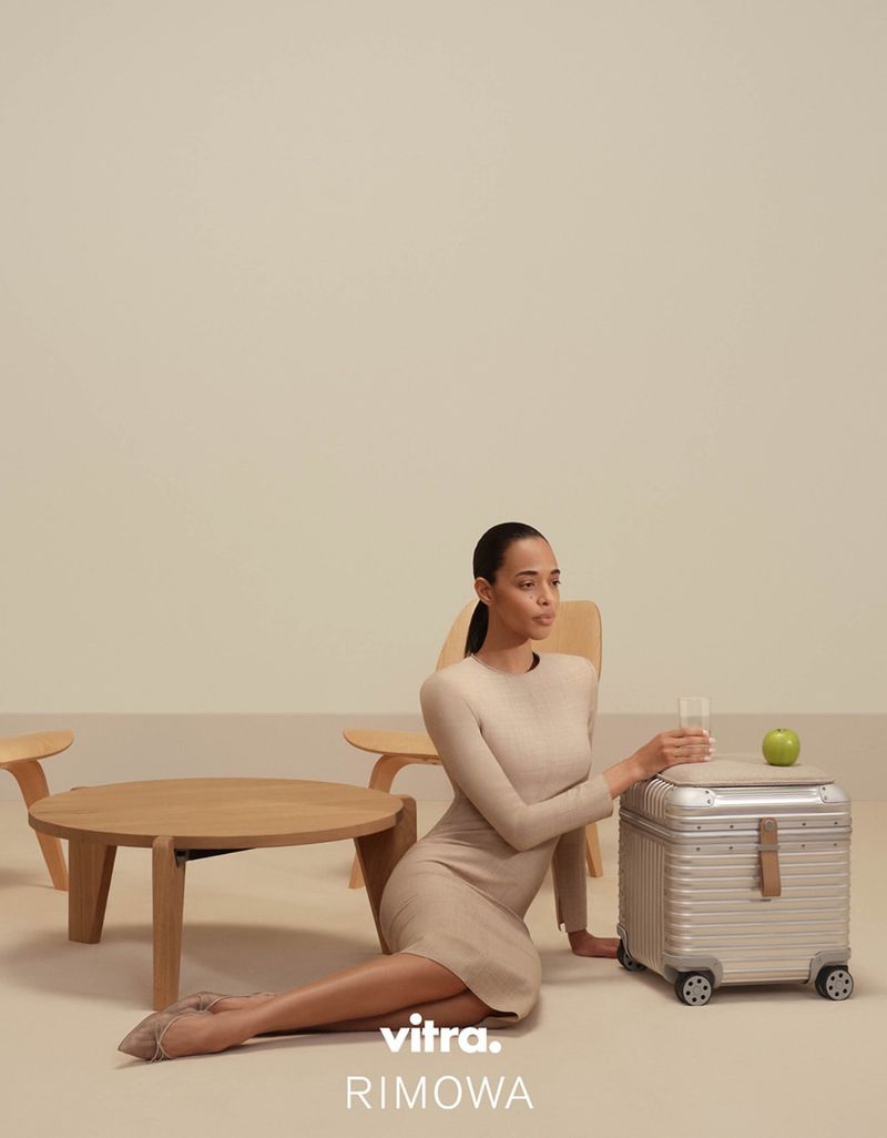 A woman in a beige dress sits next to a metallic suitcase with an apple and glass of water on top, in a neutral studio setting.