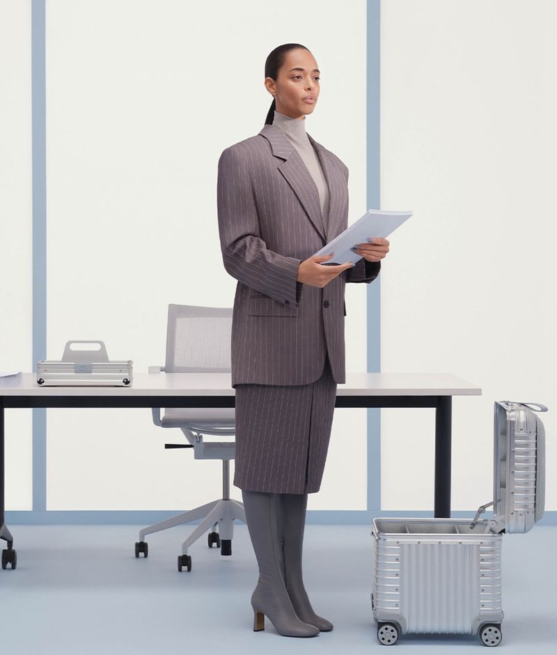 Businesswoman in pinstripe suit stands in office setting with open aluminum suitcase, holding papers. Interior shot, eye-level angle.