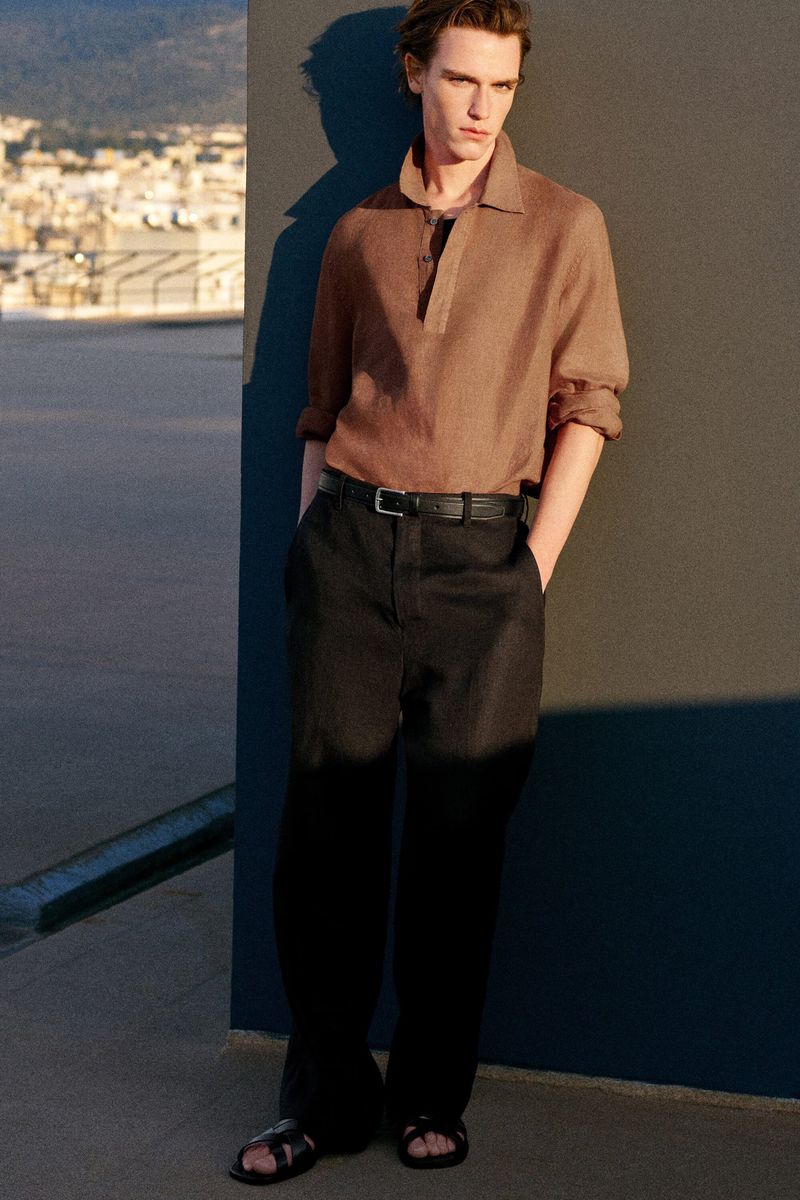 A male model leaning against a wall wearing a brown long-sleeve polo shirt and dark trousers for COS.