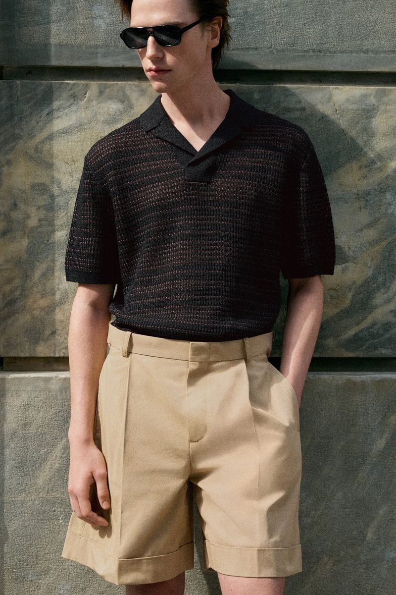 Model wearing dark sunglasses, a black knit polo, and beige shorts standing against a stone wall for COS.