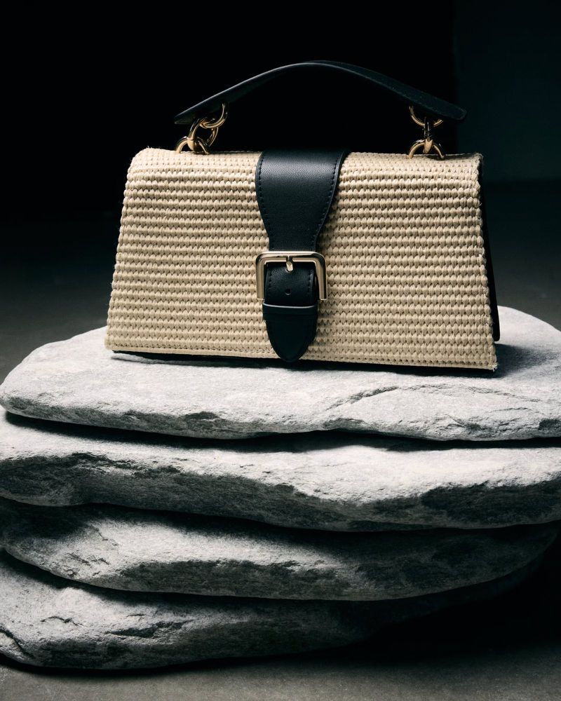 Beige woven handbag with a black leather strap and gold buckle, placed on a stack of four flat, gray stones against a dark background.