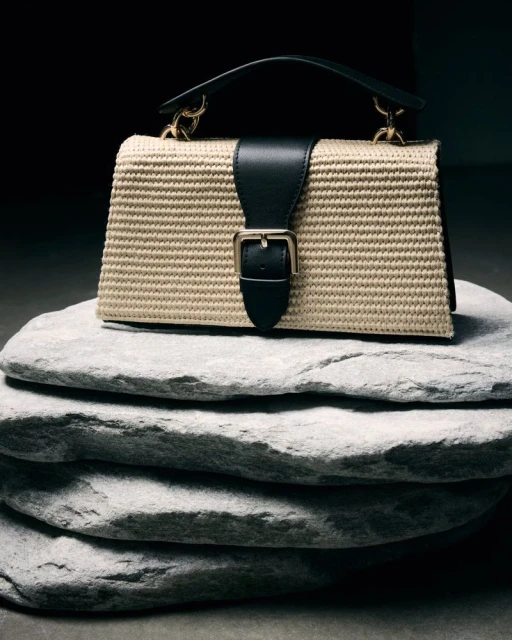 Beige woven handbag with a black leather strap and gold buckle, placed on a stack of four flat, gray stones against a dark background.