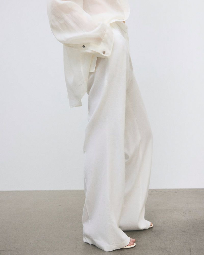 Fashionable woman in wide-leg white pants and a semi-sheer white top stands against a white wall, showcasing neutral-tone fashion.