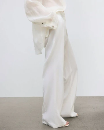 Fashionable woman in wide-leg white pants and a semi-sheer white top stands against a white wall, showcasing neutral-tone fashion.
