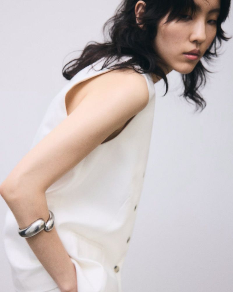 A young woman with black hair and a sleeveless white top poses indoors with a silver bracelet against a neutral background.