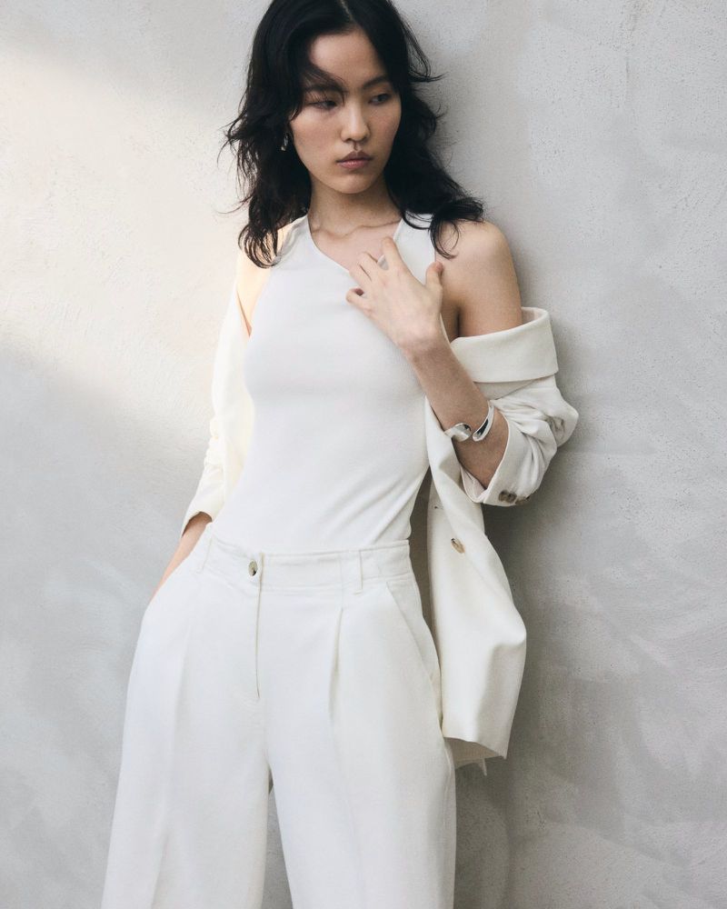 Portrait of a woman in white tank top and wide-leg pants posing against a concrete wall, with a blazer draped over her shoulder.