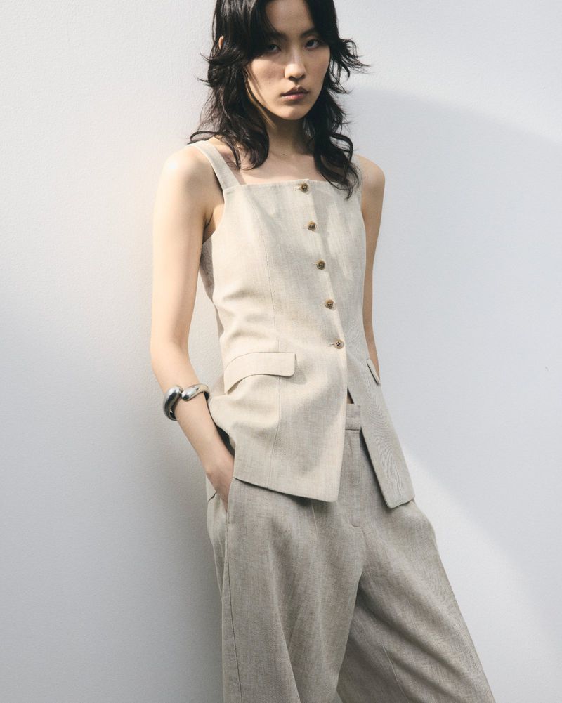 Asian woman poses in a sleeveless linen top and linen trousers against a white wall in a studio, wearing a silver bracelet.