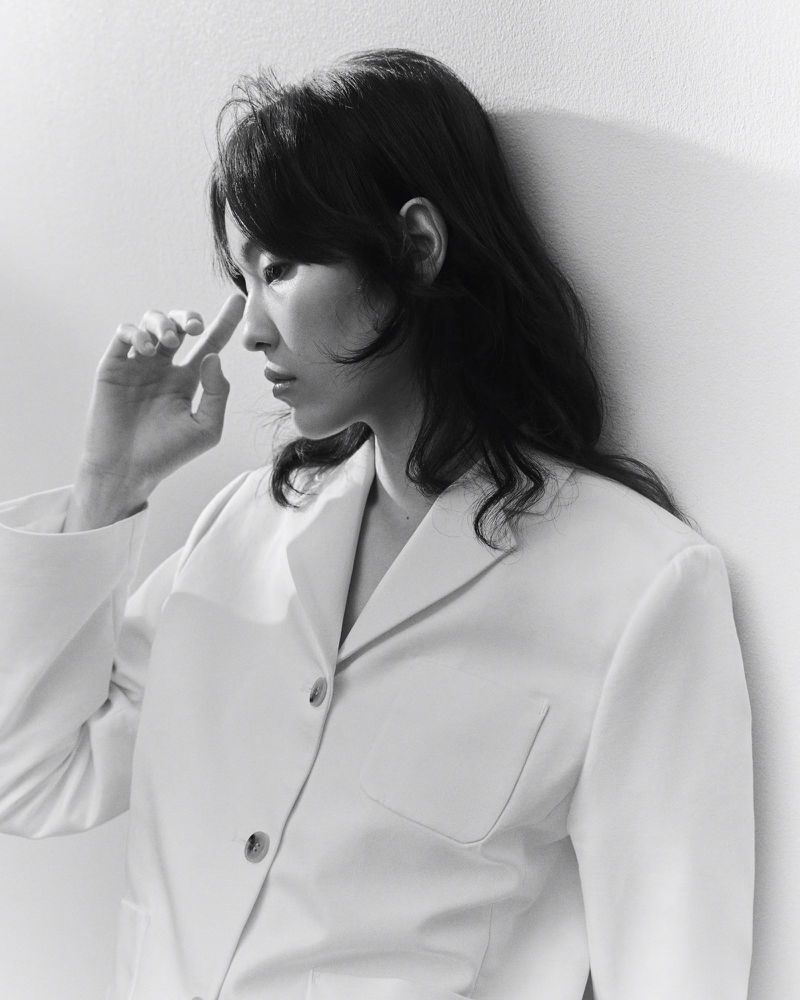 Black and white portrait of an Asian woman with dark hair, wearing a white blazer, leaning against a white wall in a contemplative pose.