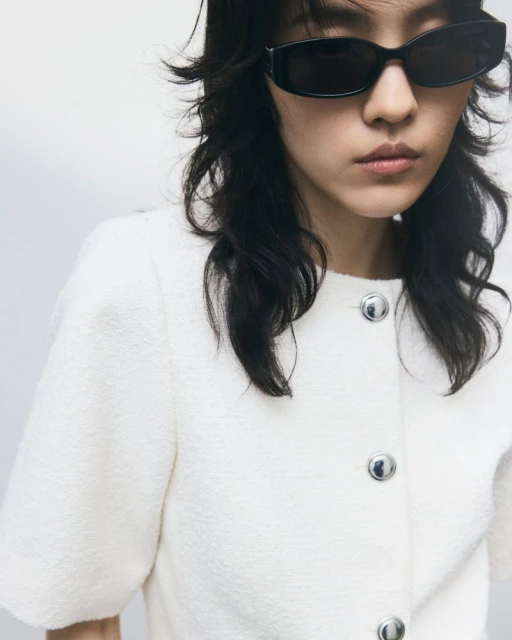 Close-up of a stylish woman with dark hair wearing black sunglasses and a white jacket with silver buttons against a white backdrop.