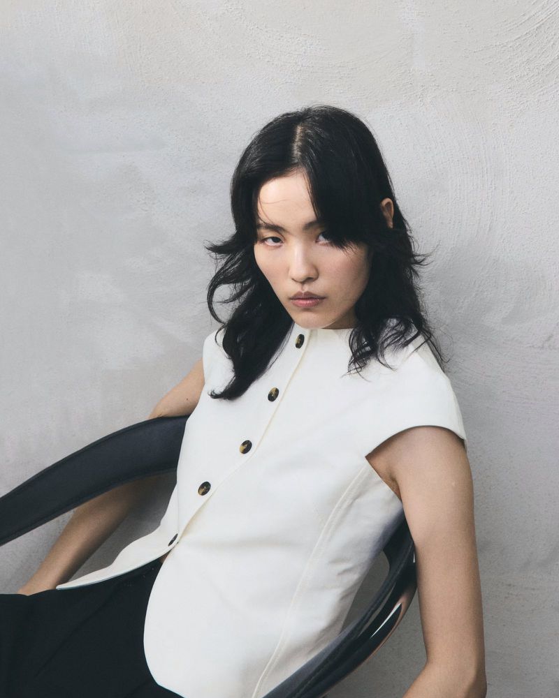 Portrait of an Asian woman with dark wavy hair wearing a white button-down shirt sitting in a black chair in front of a grey wall.