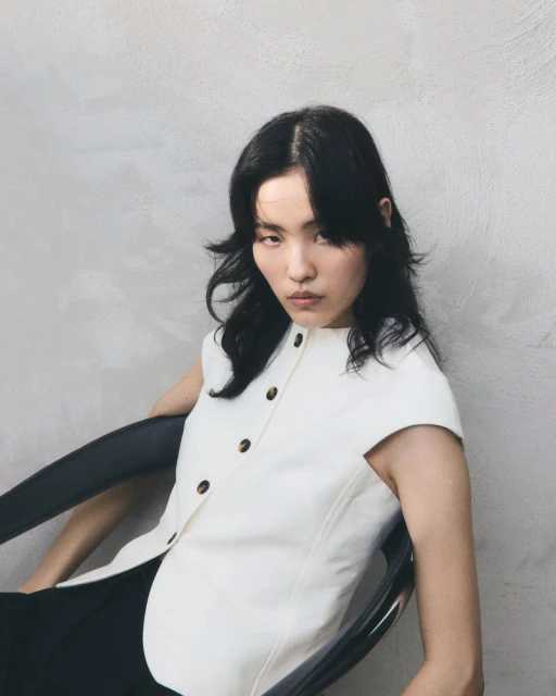 Portrait of an Asian woman with dark wavy hair wearing a white button-down shirt sitting in a black chair in front of a grey wall.