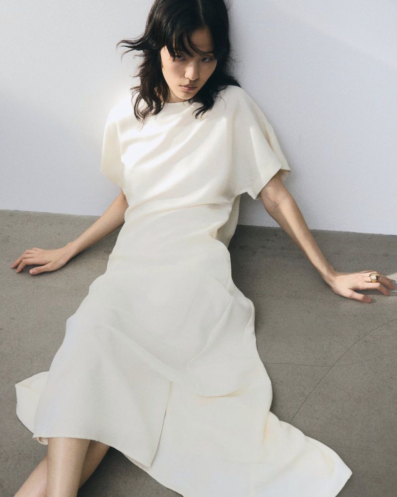 Model seated on the floor wearing a cream white dress against a plain wall