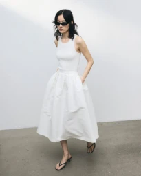 Model wearing a white tank top and voluminous skirt with black sunglasses against a white wall.