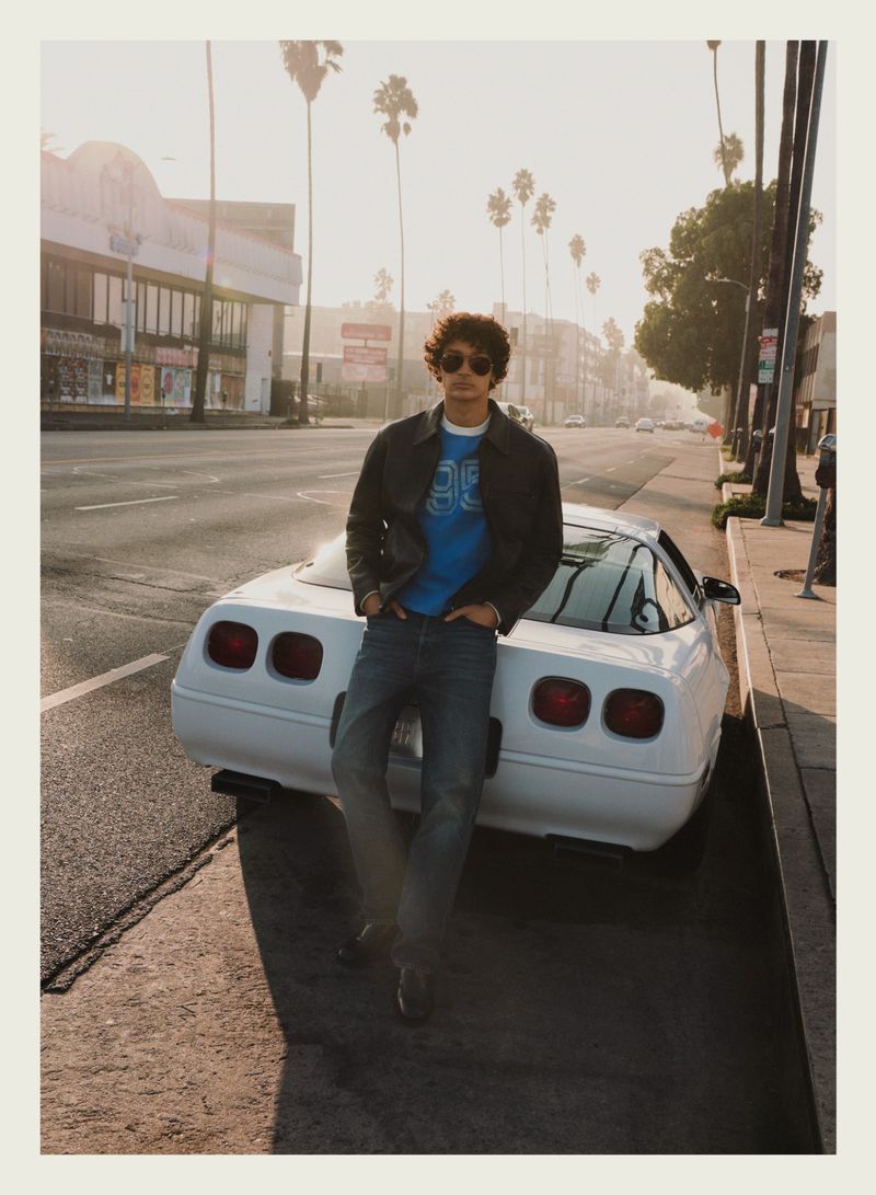 Young man wearing sunglasses and a leather jacket standing against a white sports car on a palm tree-lined street.