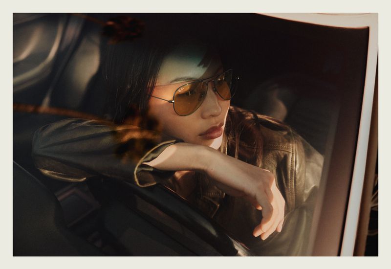 Model wearing gold-tinted aviator sunglasses and leather jacket inside car, photographed by Quentin de Briey for Ray-Ban.